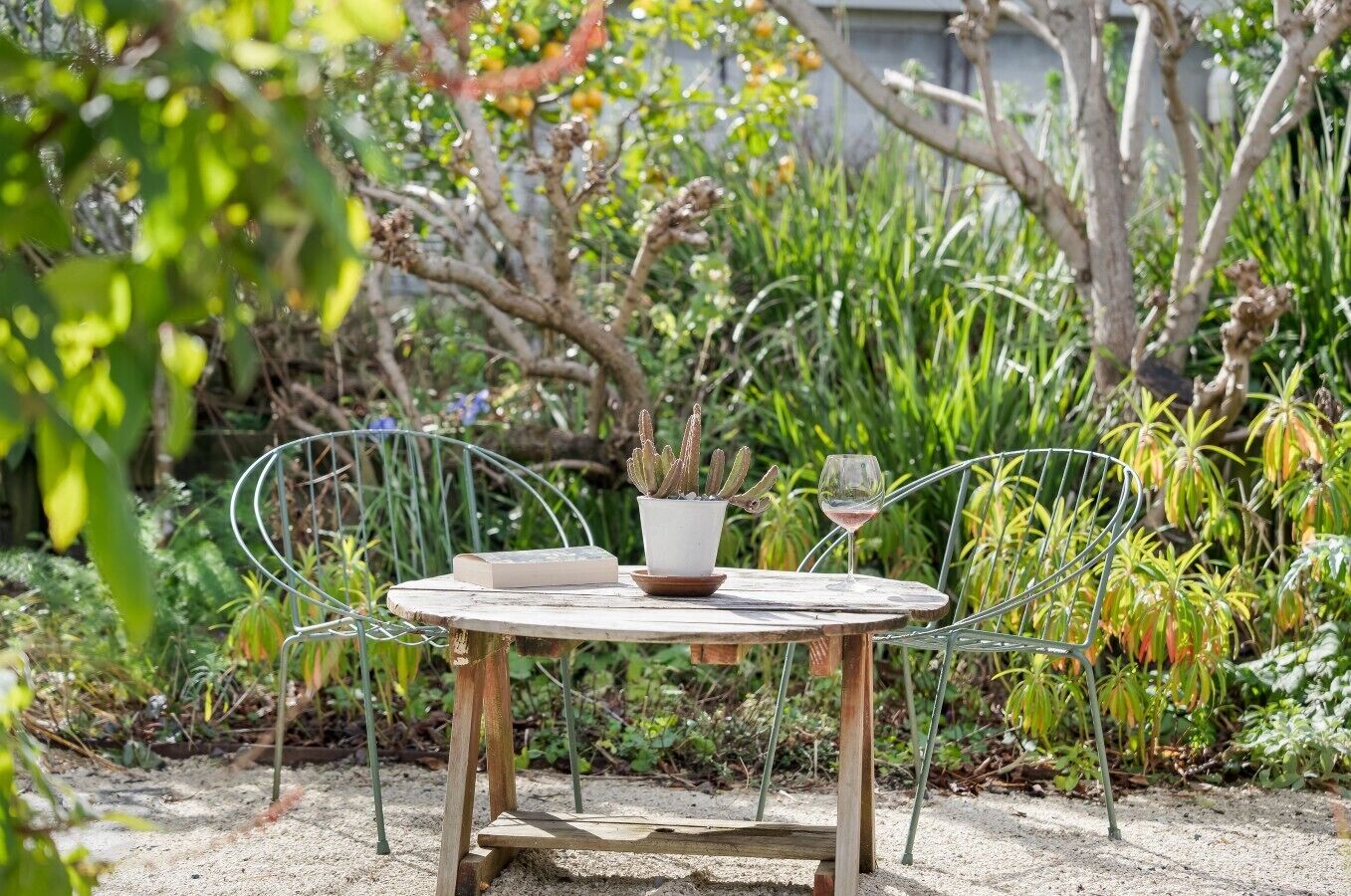 A round metal table with a white pot and a glass of wine, surrounded by greenery.