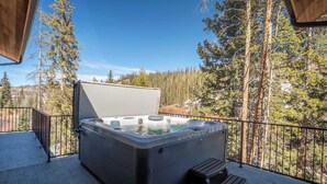 Ein Whirlpool auf einer Holzterrasse mit Metallgeländer, umgeben von Bäumen und blauem Himmel.
