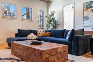 A modern living room with a dark blue sofa, a wooden coffee table, and a large window with a view of a building. - Peaceful Views | Hot Tub | Creekside Privacy (Winter Park)