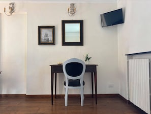 Quarto com piso de madeira, uma cadeira branca, uma pequena mesa com uma planta, um quadro emoldurado, um espelho e uma TV fixada na parede.