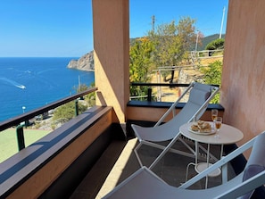Uma varanda com vista para o mar, duas cadeiras de descanso brancas, uma mesa pequena com um prato de comida e copos, e um corrimão.