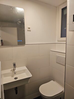 A small, clean bathroom with a white sink, a mirror, a toilet, and a window. - Zentral & Gemütliche Wohnung 2 (Köln)