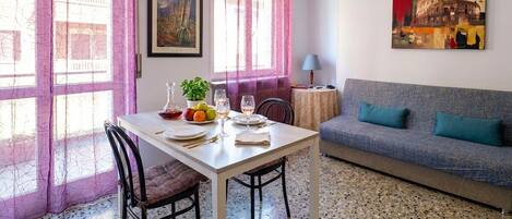 A room with a dining table set for two, a sofa, a lamp, and a painting on the wall.