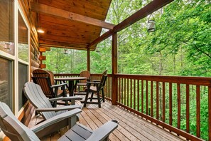 Eine Holzterrasse mit Adirondack-Stühlen und einem Tisch, mit Blick auf ein bewaldetes Gebiet.