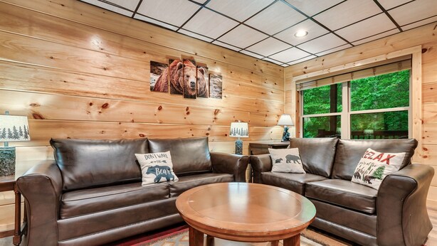 Une pièce aux murs lambrissés, avec deux canapés en cuir, une table ronde en bois et une grande fenêtre donnant sur la forêt.