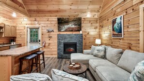 L’intérieur d’une cabane comprend une cheminée en pierre, des murs en bois, un coin cuisine et un salon avec un canapé et des tabourets de bar.