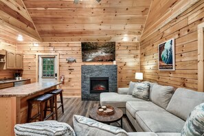 Ein Interieur einer Hütte mit Kamin aus Stein, Holzwänden, einer Küchenzeile und einem Wohnbereich mit Sofa und Barhockern.