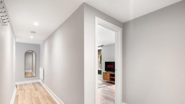 Un pasillo con piso de madera, una puerta blanca que da a una habitación con televisión y un radiador en la pared.