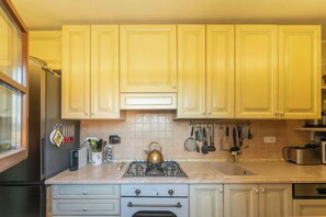 A kitchen with light wooden cabinets, a stainless steel refrigerator, a gas stove, and various kitchen utensils hanging on the wall.
