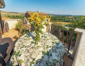Un balcón con una mesa para dos, un jarrón con girasoles y una vista panorámica del paisaje rural.