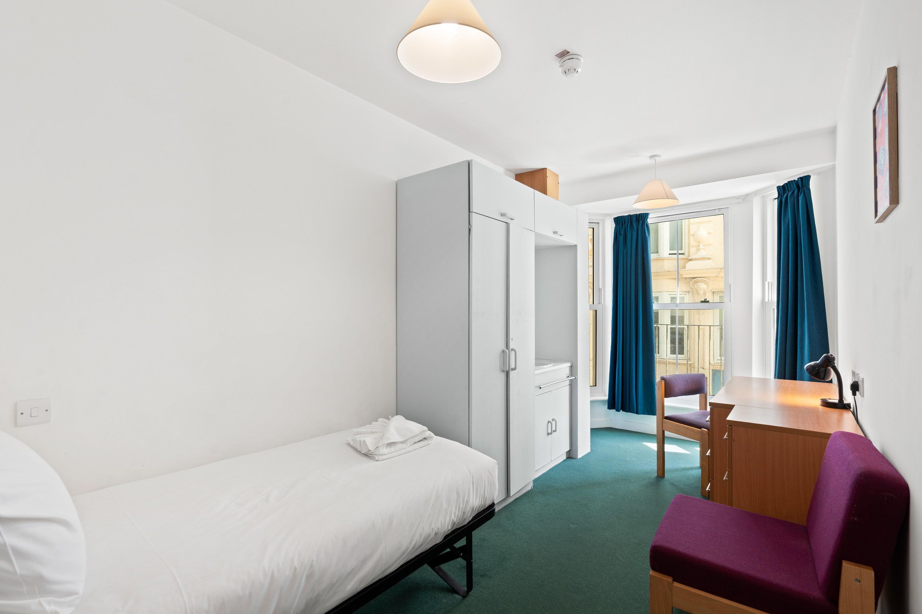 A hotel room with a single bed, a desk with a chair, a wardrobe, and a view of the outside through a window with curtains.