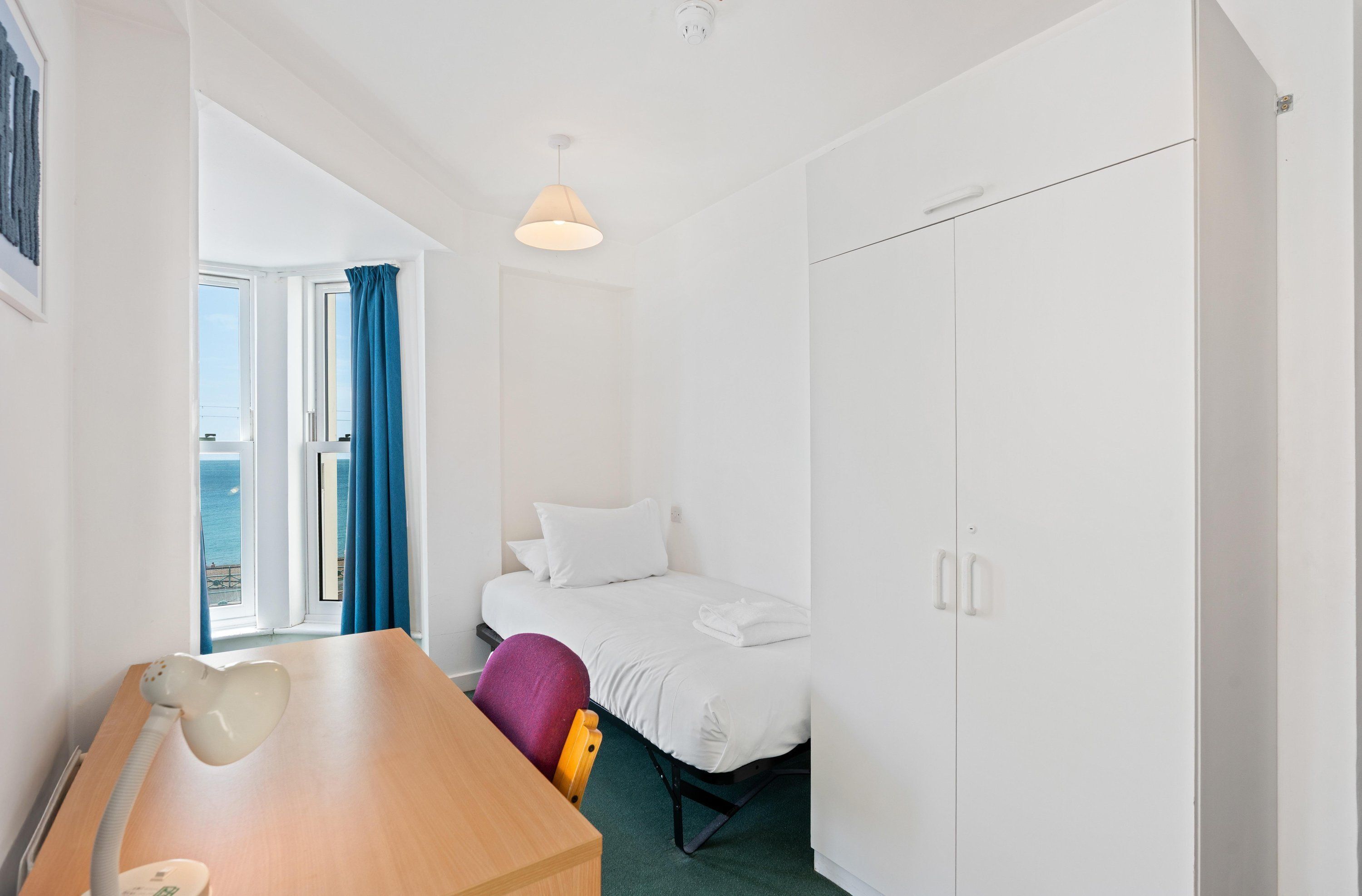 A hotel room with a bed, a desk, a chair, a wardrobe, and a view of the sea.
