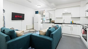 Ett modernt kök med vita skåp, en inbyggd ugn och en diskho. På väggen sitter en platt-tv och på skärmen visas ordet ”NETFLIX”.