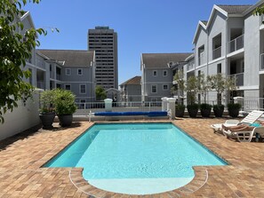 Uma piscina com deck de tijolos, rodeada por plantas em vasos e espreguiçadeiras.
