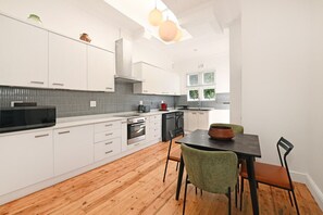 A modern kitchen with white cabinetry, a black microwave, and a wooden dining table with chairs. - Two Bed Home | Fresnaye | Secure | Spacious | BBQ (Cape Town)