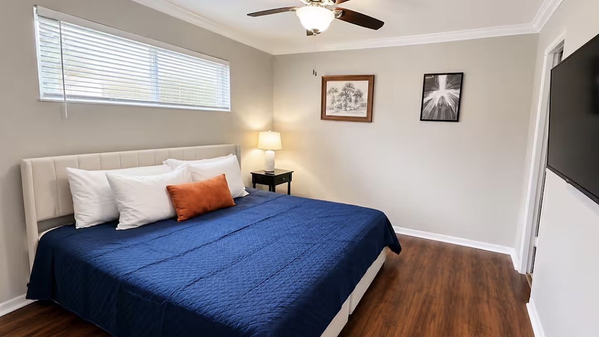 A bedroom with a bed, a ceiling fan, a nightstand with a lamp, and two framed pictures on the wall.