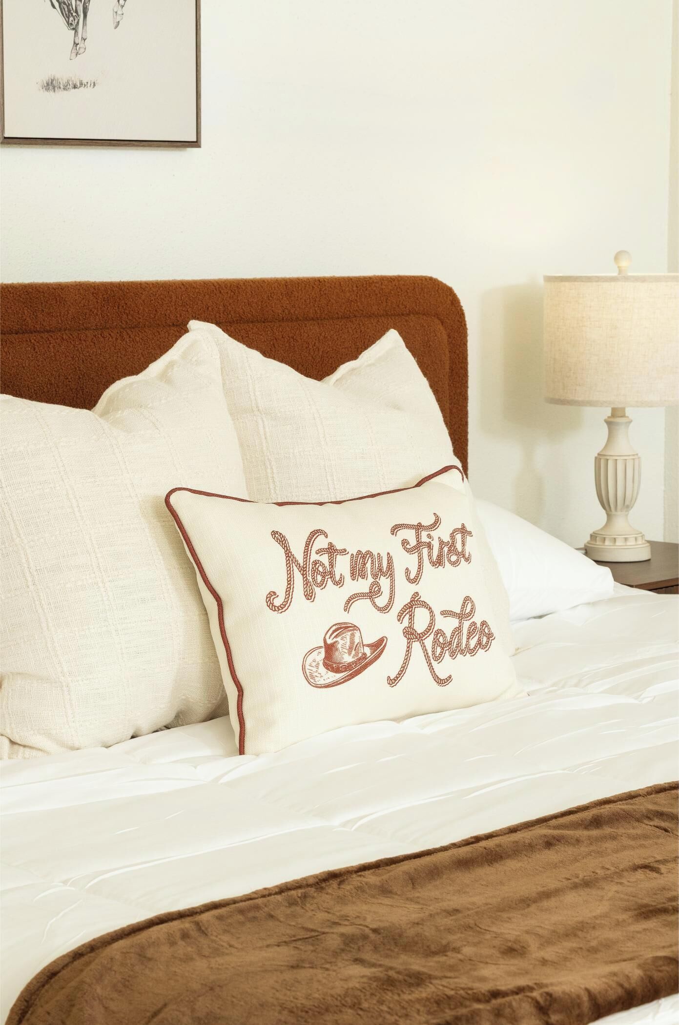 A bed with white bedding and a brown headboard, featuring a pillow with embroidered text.