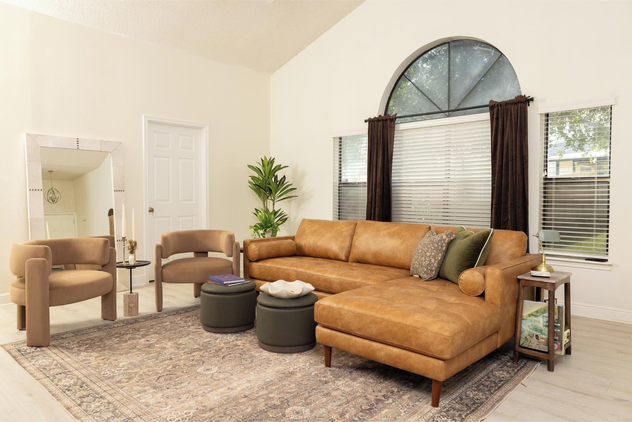 A living room with a brown leather sofa, two armchairs, a coffee table, a potted plant, and a large window with blinds.