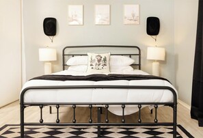 Una cama con estructura metálica, ropa de cama blanca, una colcha de color negro y un tapete estampado. Hay lámparas de pared y cuadros enmarcados sobre la cama.