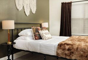 Un letto con lenzuola bianche, una coperta marrone in pelo e cuscini decorati. C'è una lampada da comodino su un supporto in metallo nero, e una decorazione in macramè appesa al muro.