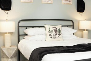 Una cama bien hecha con sábanas blancas, un plaid negro y una almohada decorativa con un paisaje desértico. Hay dos lámparas de noche sobre mesas blancas, y la habitación tiene un diseño sencillo y limpio con una pared de color claro.