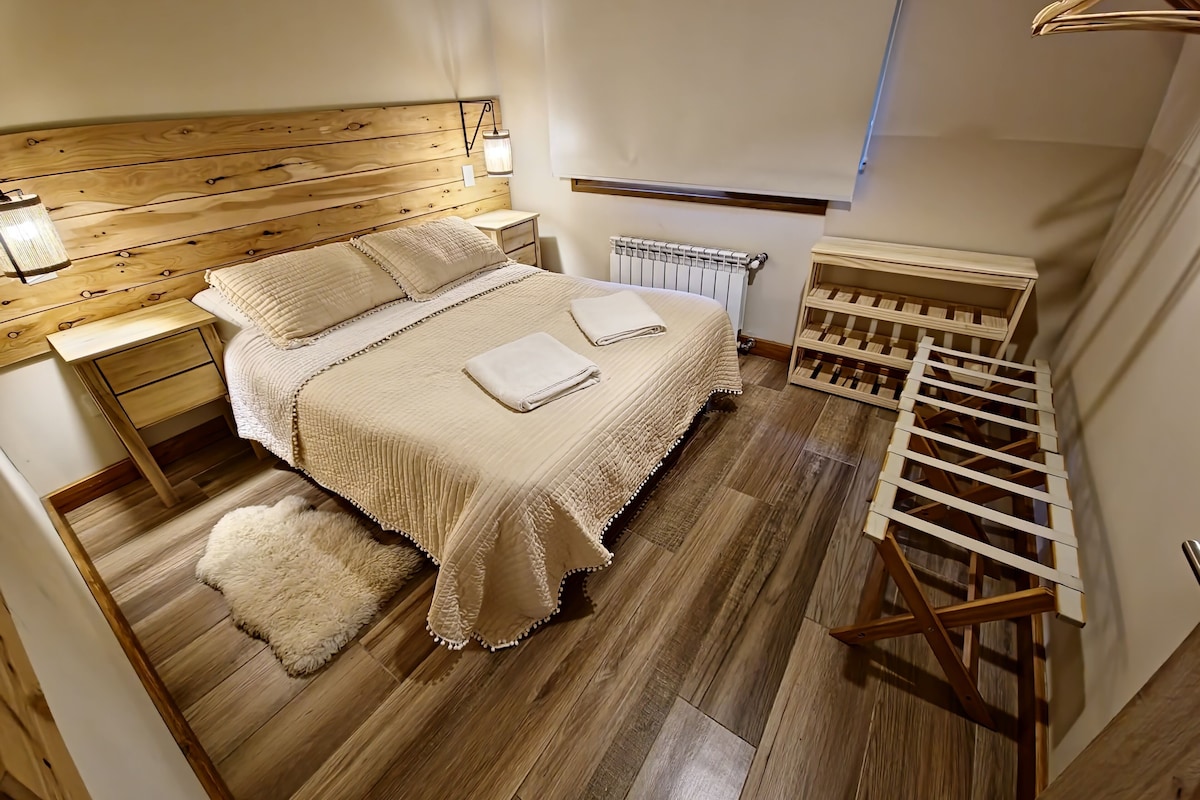 Ein Schlafzimmer mit hölzernem Kopfteil, einem Nachttisch, einem Heizkörper, einer Pendelleuchte, Holzboden und einem eingebauten Weinkühlschrank.