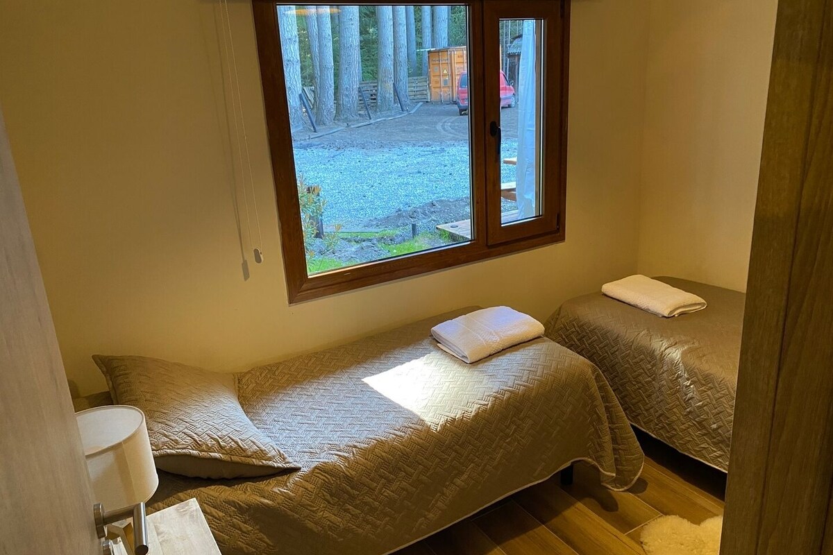 Ein kleiner, gemütlicher Raum mit zwei Einzelbetten, einem Fenster mit Blick auf einen Wald und einen Kiesweg, sowie einem Holzboden.