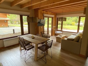 Quarto espaçoso com teto de madeira, mesa de jantar com cadeiras, sofá e grandes janelas com vista para o exterior.