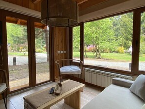 A room with a large window, a hanging light fixture, a white sofa, a wooden table, and a chair. - Cabañas en el bosque patagónico (Villa La Angostura)