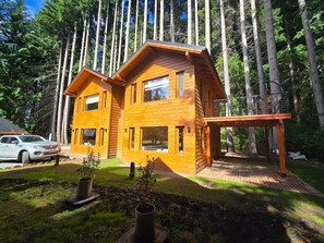 A two-story wooden cabin with a deck, surrounded by tall trees. - Cabañas en el bosque patagónico (Villa La Angostura)