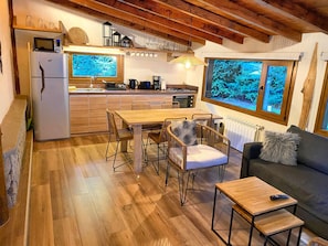 Cozinha compacta com geladeira, micro-ondas e fogão, área de jantar com mesa e cadeiras, e sala de estar com sofá e janela com vista para o verde.