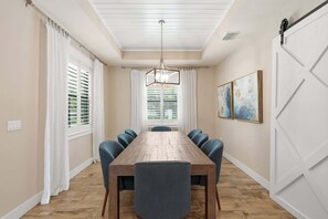 Une salle à manger avec une table en bois, six chaises, un lustre et de grandes fenêtres ornées de rideaux blancs.