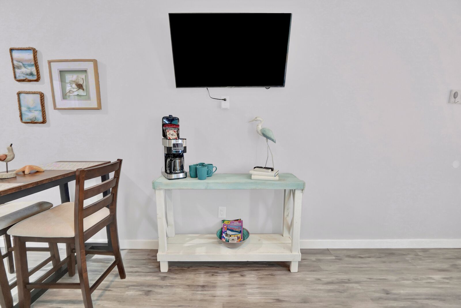 A room with a wooden dining table, a coffee maker, a teal console table with a bird sculpture, and a television mounted on the wall.