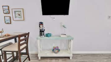 A room with a wooden dining table, a coffee maker, a teal console table with a bird sculpture, and a television mounted on the wall.