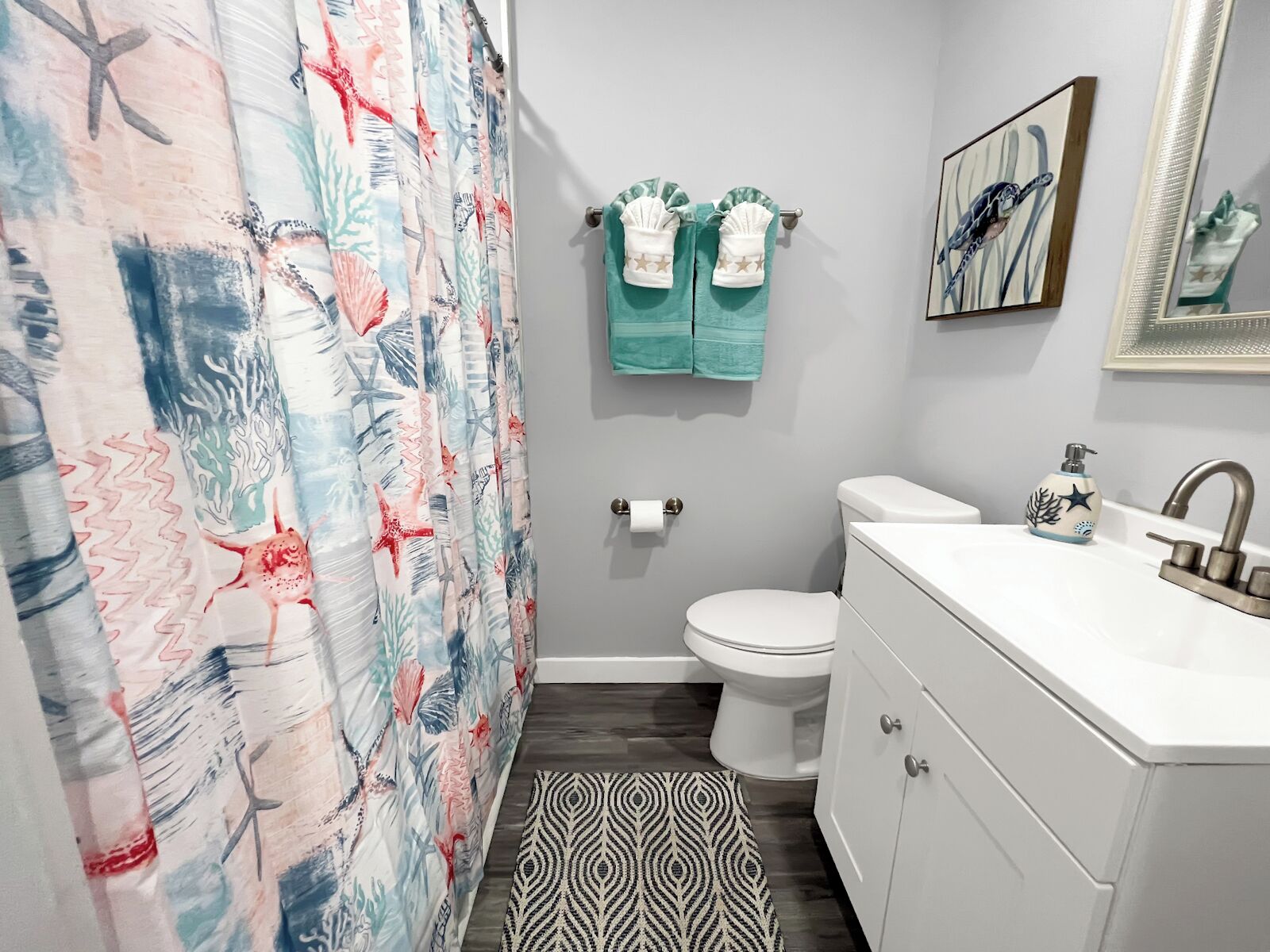 Un baño con cortina de ducha de diseño marino, inodoro, lavabo con dispensador de jabón decorativo y alfombra estampada en el suelo.