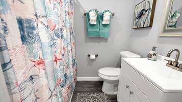 Un baño con cortina de ducha de diseño marino, inodoro, lavabo con dispensador de jabón decorativo y alfombra estampada en el suelo.