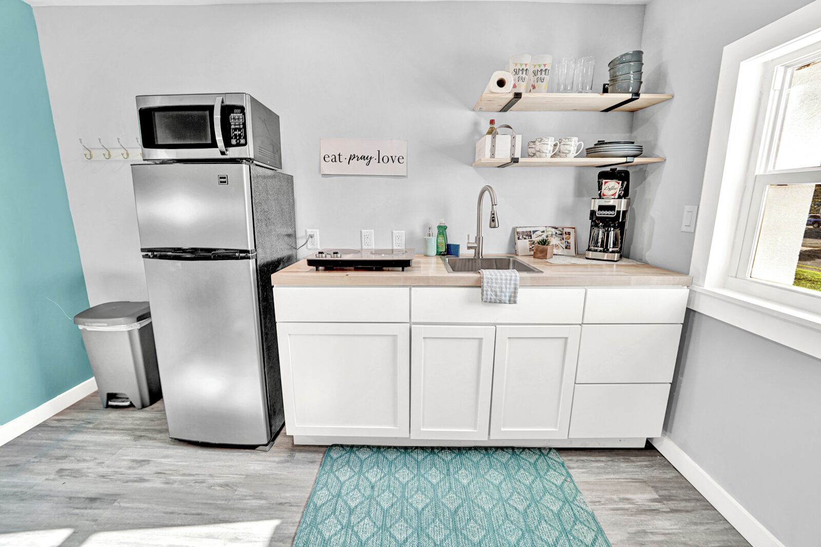A compact kitchen with a stainless steel refrigerator, a microwave, a sink, and a coffee maker. There is a small trash bin and a decorative rug.