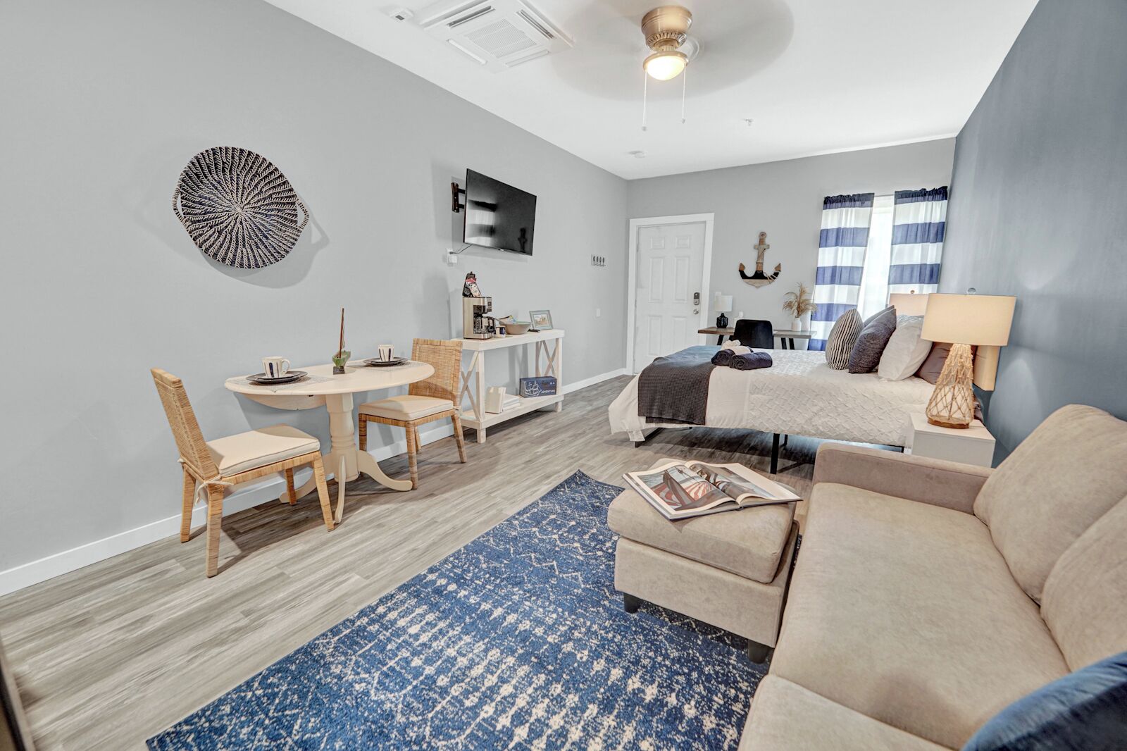 A modern living room with a sofa, a dining table with chairs, a TV mounted on the wall, and a ceiling fan.