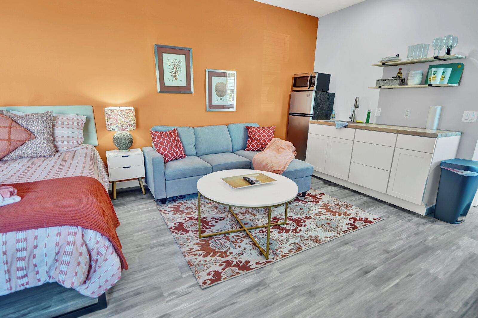 A modern bedroom with a bed, a sofa, a small round table, and a kitchenette area with a microwave and refrigerator.