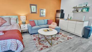 A modern bedroom with a bed, a sofa, a small round table, and a kitchenette area with a microwave and refrigerator.