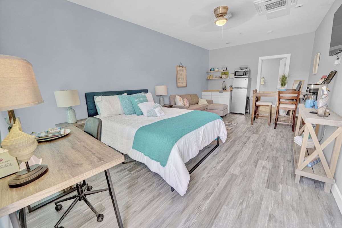 A bedroom with a bed, bedside tables, a desk, and a kitchenette.