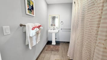 Une salle de bain avec un lavabo blanc, un miroir, un porte-serviettes avec des serviettes, un rideau de douche et un plancher en bois.