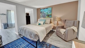 A modern hotel room with a bed, a chair, a desk, and a painting on the wall.
