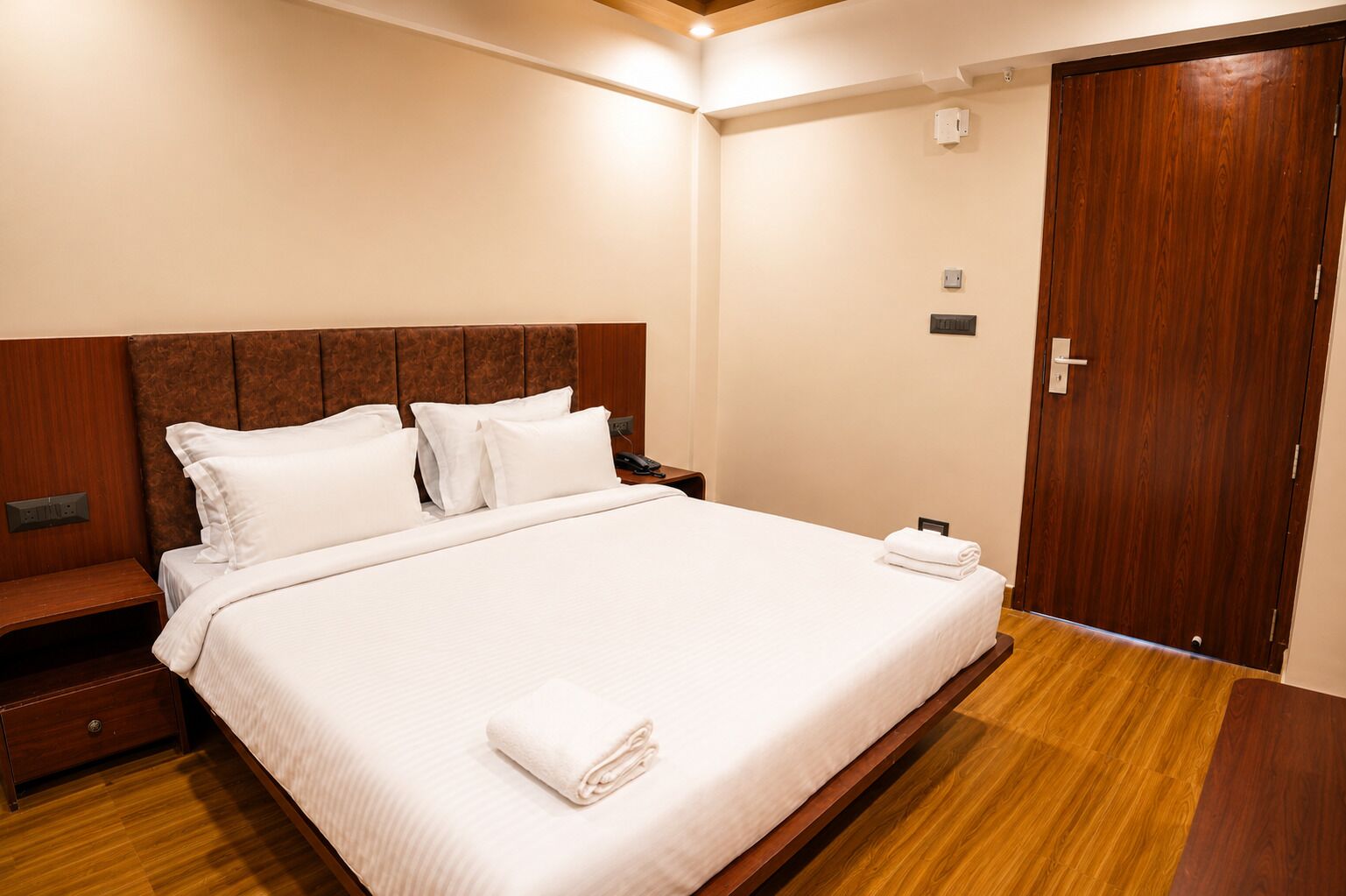 A hotel room with a large bed, wooden headboard, a wooden nightstand, a wooden wardrobe, and a wooden floor.