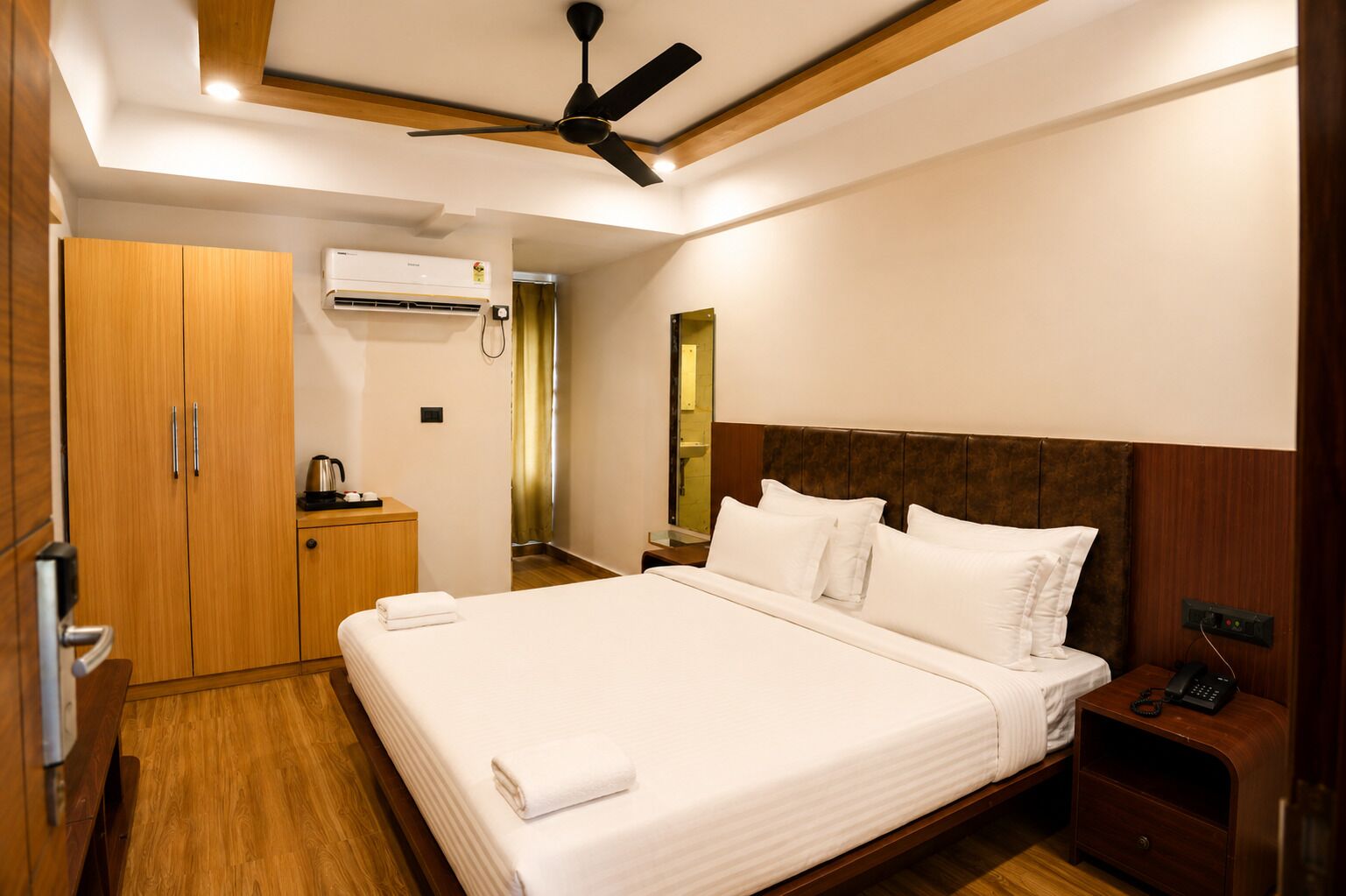 A hotel room with a bed, bedside table, nightstand, wardrobe, and an air conditioning unit.