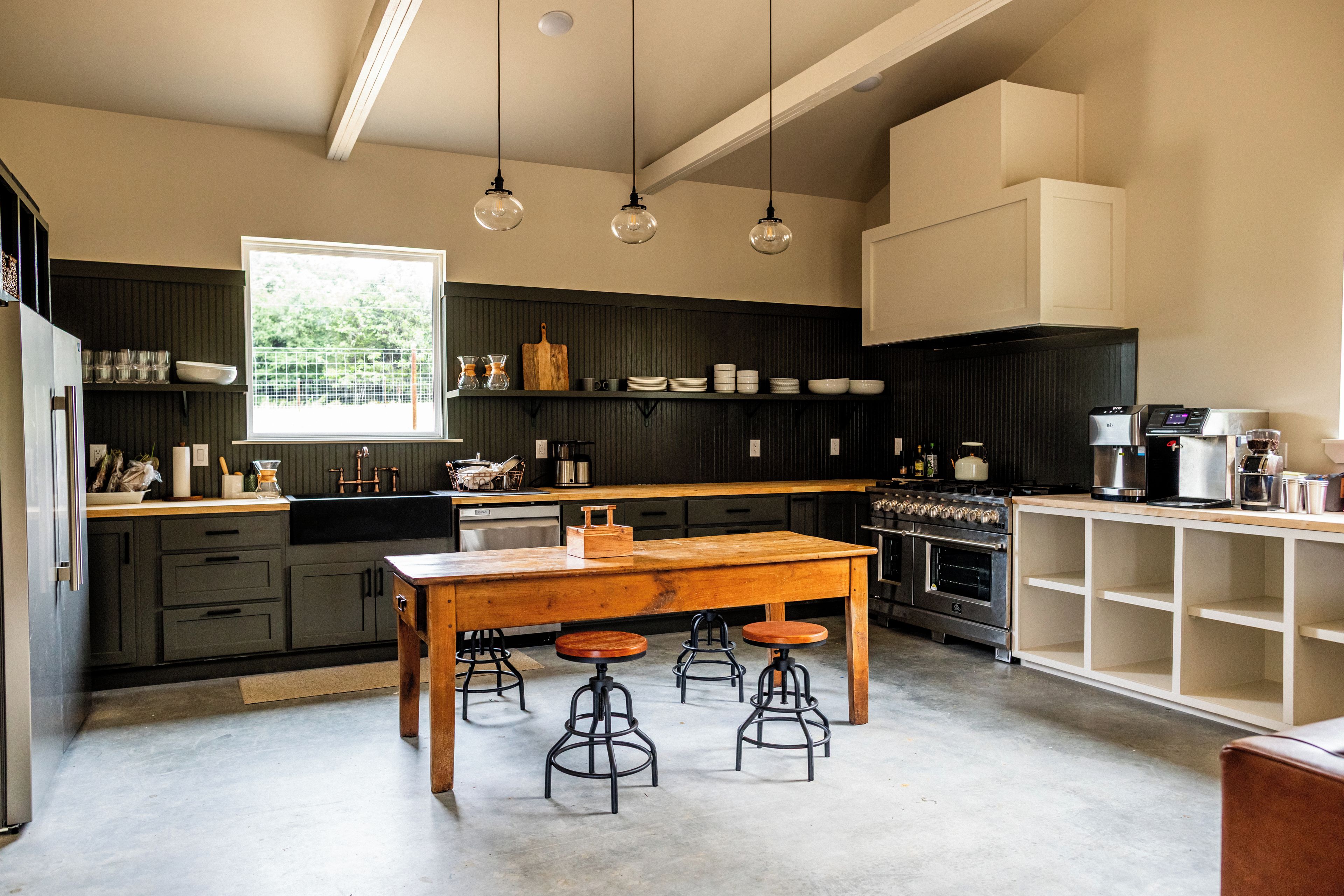 A modern kitchen with dark cabinetry, a wooden table, bar stools, and a large window.
