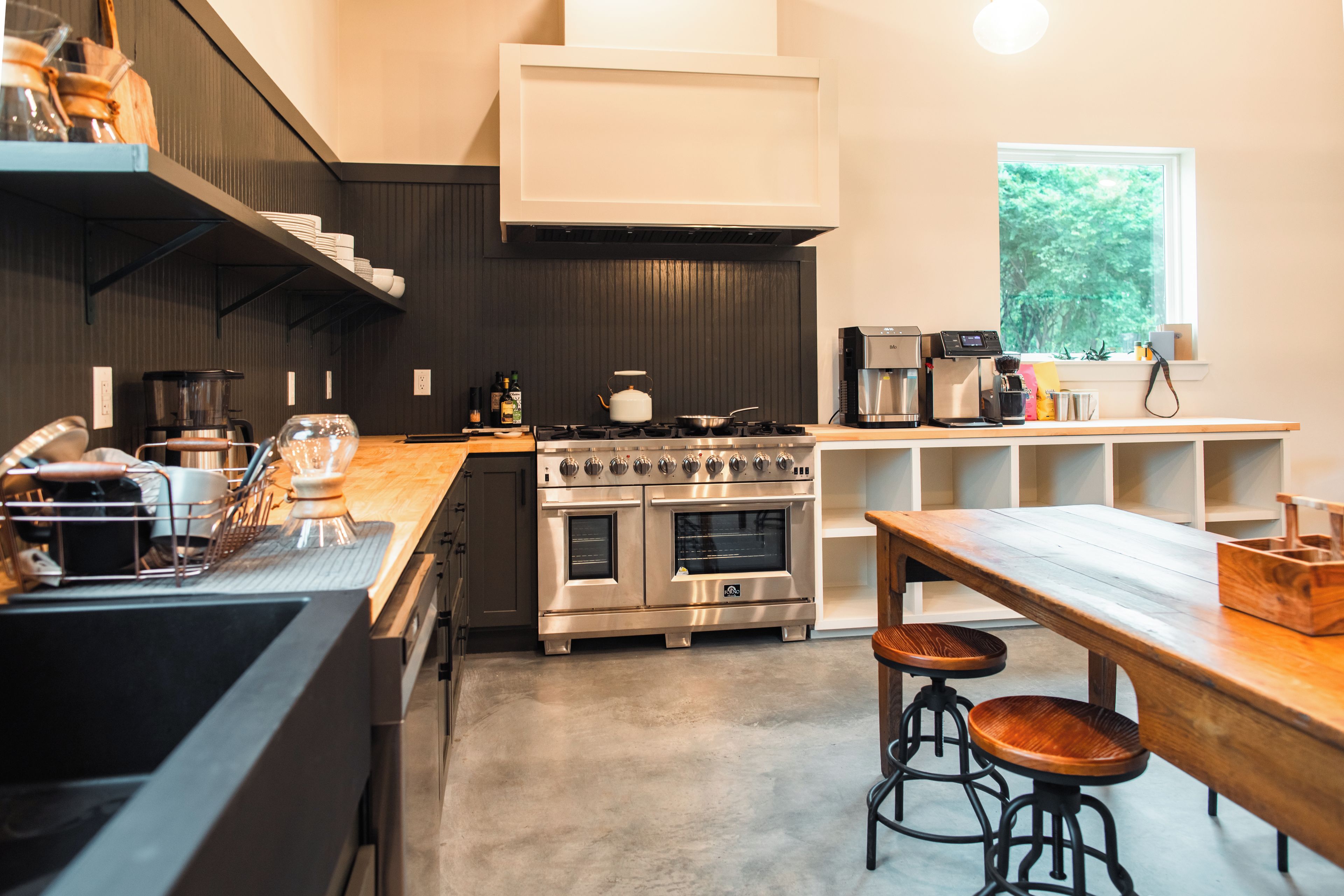 A modern kitchen with a large stove, oven, and coffee machine. There are stools and a wooden table.