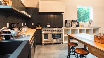 A modern kitchen with a large stove, oven, and coffee machine. There are stools and a wooden table.