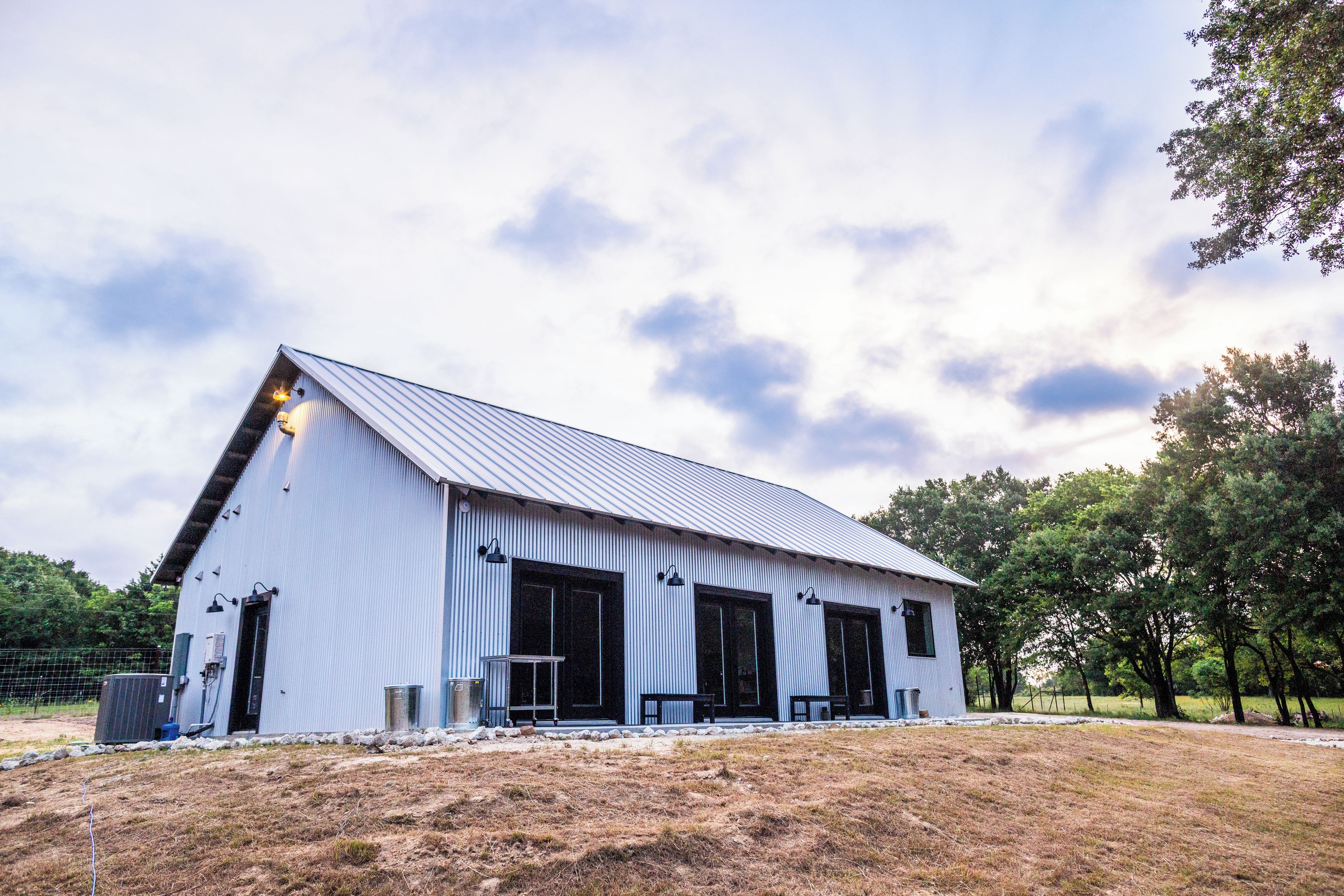 A large, single-story building with a sloped roof, likely a barn or storage facility, surrounded by a grassy area and trees.