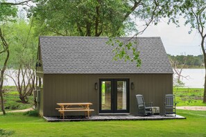 Une petite cabane avec un toit sombre, une porte en bois et une table de pique-nique à l’extérieur.